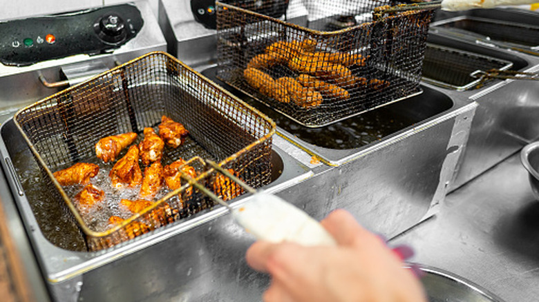 A hand holding a fryer basket full of chicken wings in a small commercial deep fryer