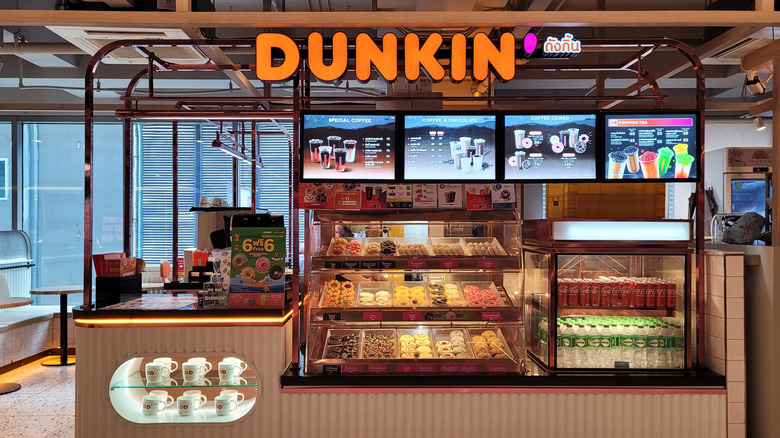 The exterior of a Dunkin' kiosk, with donuts, drinks, menus, and mugs displayed prominently