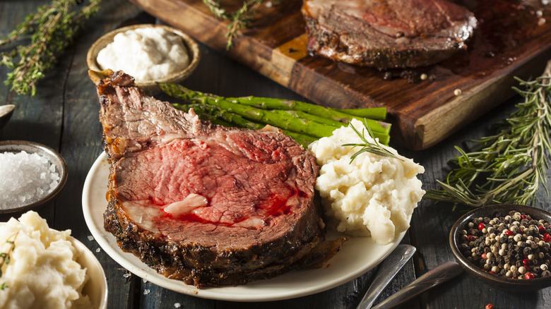 A meal of prime rib and asparagus with mashed potatoes surrounded by salt, peppercorns, and herbs