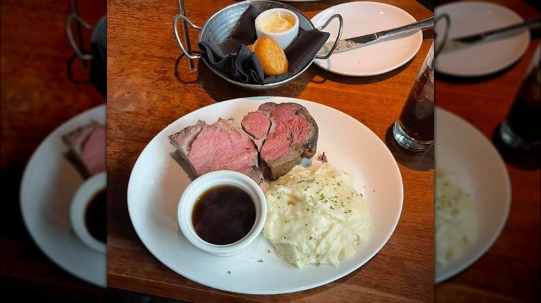 A piece of prime rib with mashed potatoes and au jus on a white plate next to a basket of dinner rolls