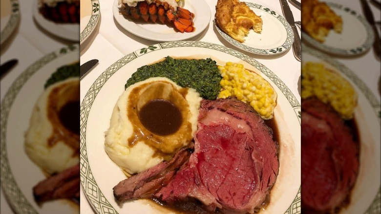 A plate of rare prime rib, potatoes, and creamed vegetables with lobster and bread in the background on a restaurant table