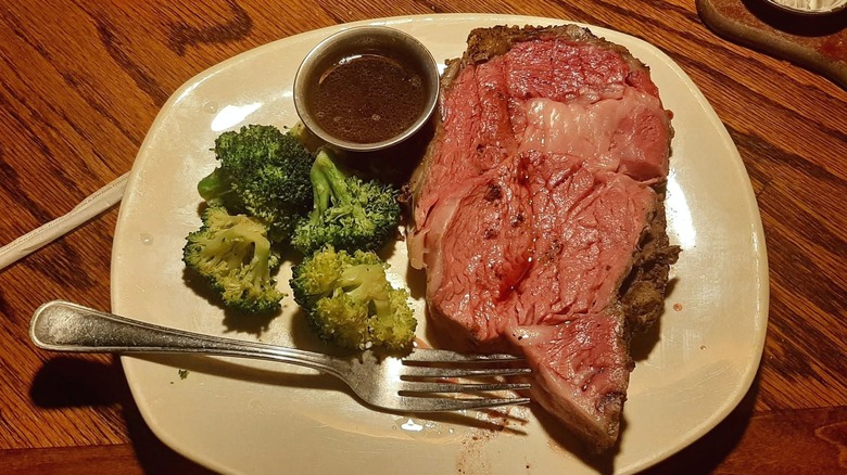 The herb-crusted prime rib meal at Outback Steakhouse with broccoli and a ramekin of sauce on a white plate