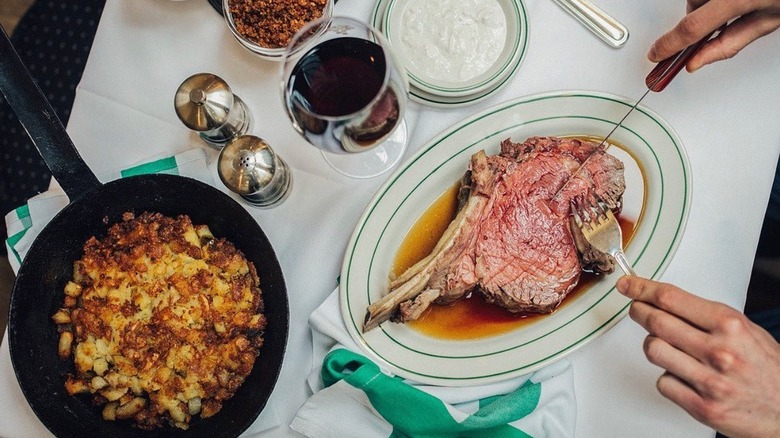 A hand cuts into a prime rib dinner with a potato skillet and glass of wine set on a white tablecloth