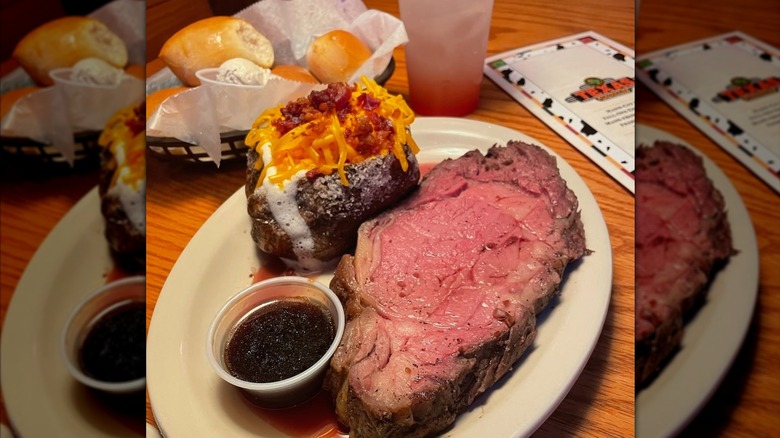 A Texas Roadhouse prime rib and a potato on a white plate with bread rolls behind it