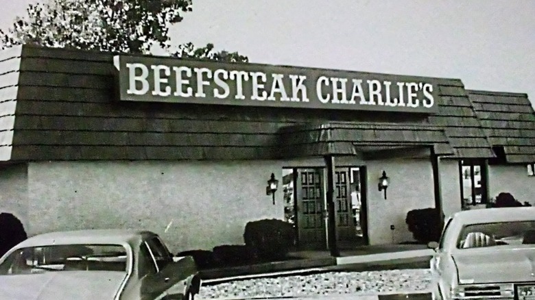 A black-and-white image of a Beefsteak Charlie's restaurant, with its sign visible and several cars