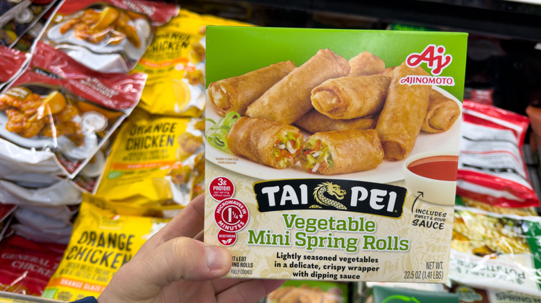 A person holding a box of frozen spring rolls with a variety of frozen Chinese food in the background
