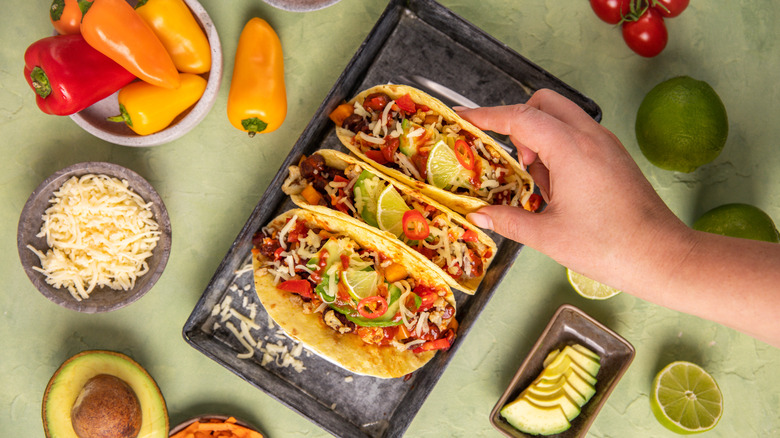 A hand takes a taco from a plate of three surrounded by accoutrements
