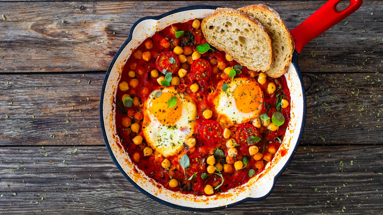 A skillet full of shakshouka with beans and toast on a wooden table