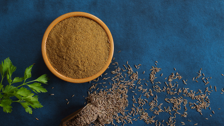 Cumin powder in a small wood bowl with seeds, on a blue background