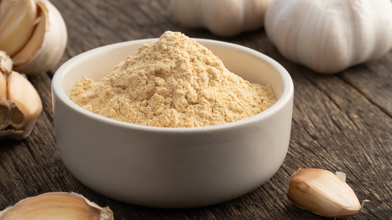 A small white bowl of garlic powder surrounded by whole garlic bulbs