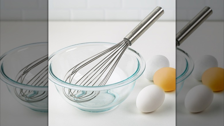 A metal balloon whisk in clear bowl next to eggs on a white countertop.