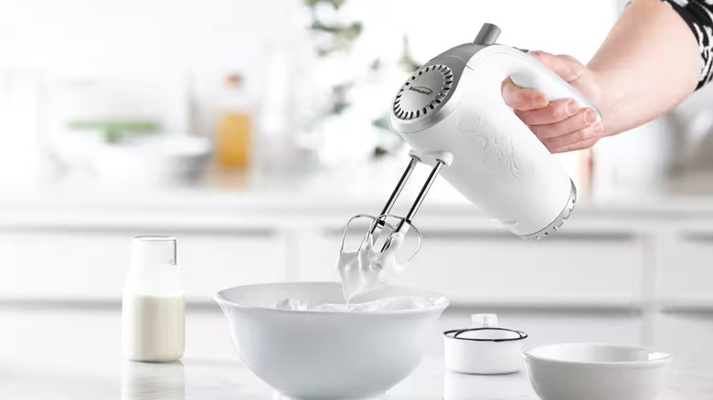 Hand holding white mixer over whipped egg whites in a white bowl on a white countertop.