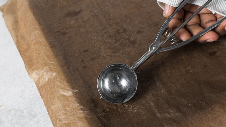Hand holding metal cookie scoop over parchment-lined cookie sheet