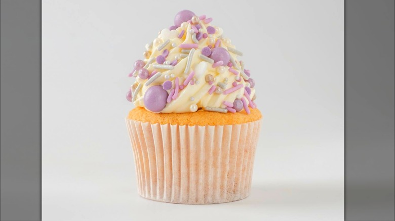Cupcake with white frosting and purple/gray sprinkles against lightly colored background.