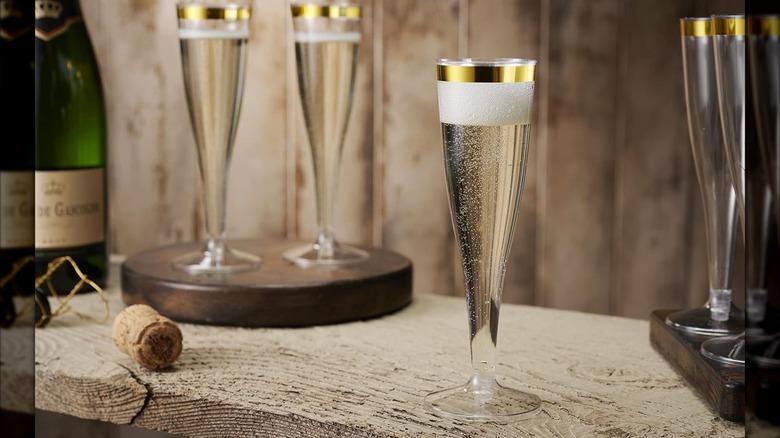 Product image of Munfix Gold Rimmed Plastic Champagne Flutes on a table filled and next to a bottle of Champagne