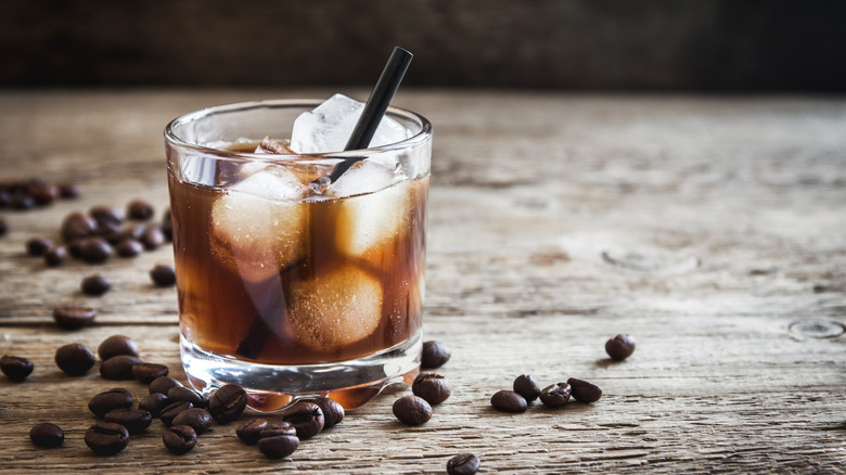 A glass of black Russian cocktail with ice and straw and coffee beans on wood