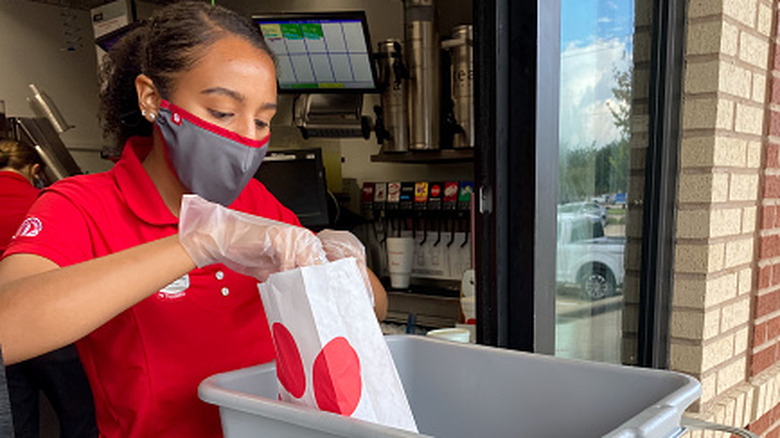 A Chick-fil-A employee in a mask packing up a customer order at the drive-thru window
