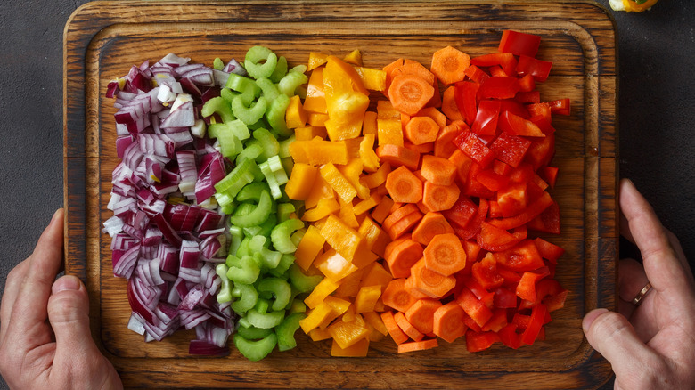 Hands holding a cutting board of various chopped vegetables lined up by color on a wooden cutting board.