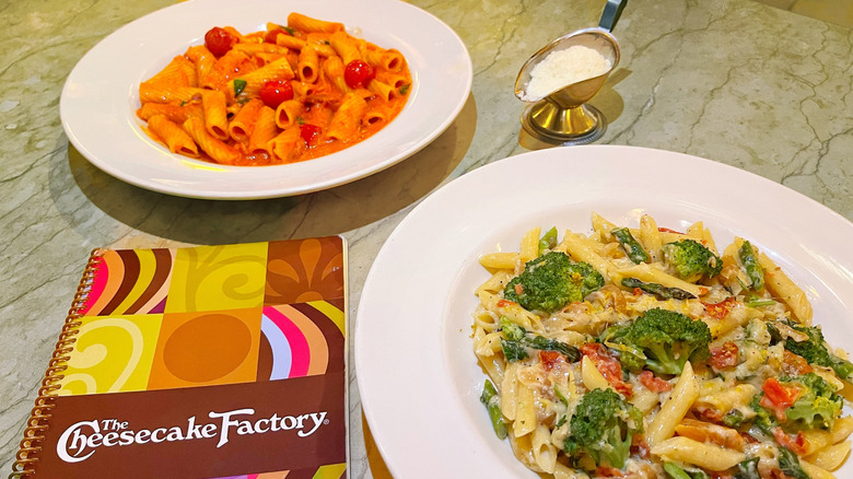 Two plates of pasta next to a Cheesecake Factory menu and a side of Parmesan cheese