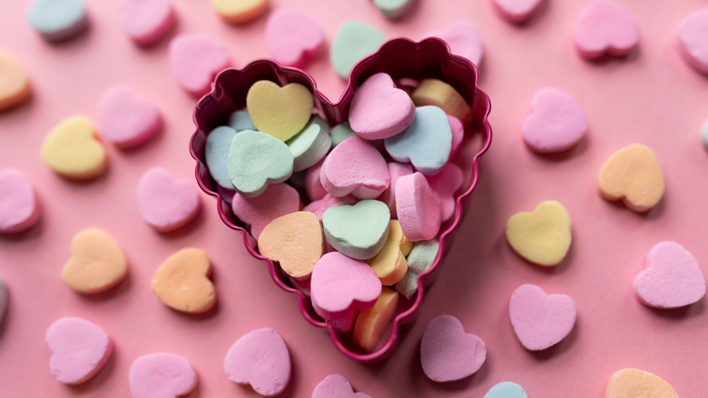 Generic candy hearts inside a heart-shaped cookie cutter against a pink background.