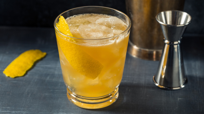 Gold rush cocktail, garnished with lemon peel, next to bartending equipment