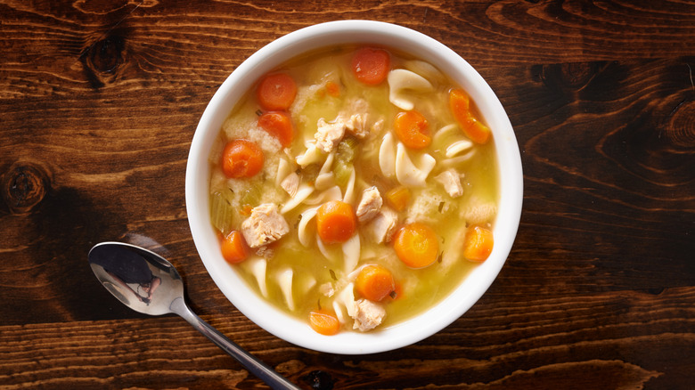 A bowl of chicken noodle soup with noodles, chicken, and carrots on a wood table