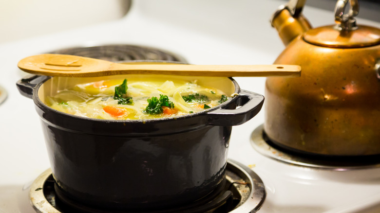 Chicken noodle soup in a black Dutch oven on an electric range with a wooden spoon over the top of the pot