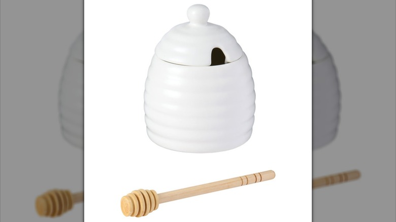 A white honey pot shaped like a beehive with a wooden honey wand against a white backdrop.