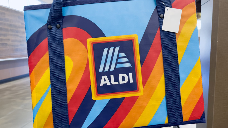 A reusable Aldi shopping bag with navy, red, orange, and yellow curves on a light blue background with an Aldi logo in the middle and navy canvas straps.