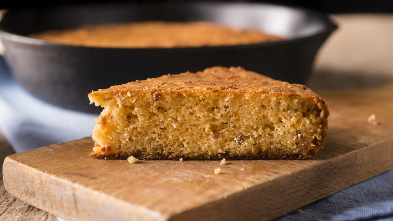 A slice of cornbread is on a cutting board with a skillet in the background.