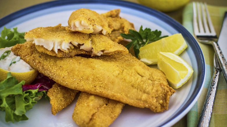 A plate of Southern-style fried fish with lemon wedges and greens is on a table.