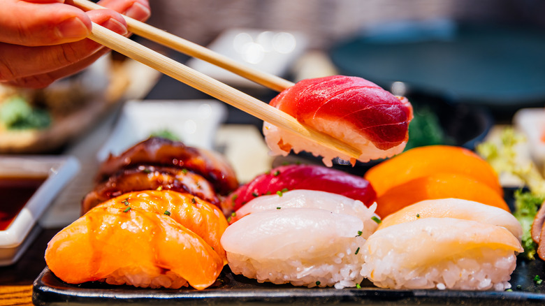 A hand lifts a piece of sushi from a platter with chopsticks