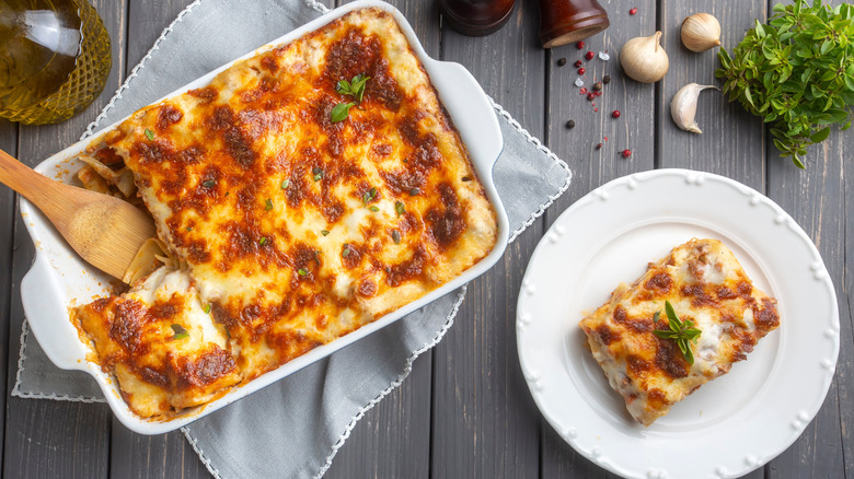 A piece of lasagna on a white plate next to a baking dish full of lasagna on a wooden table