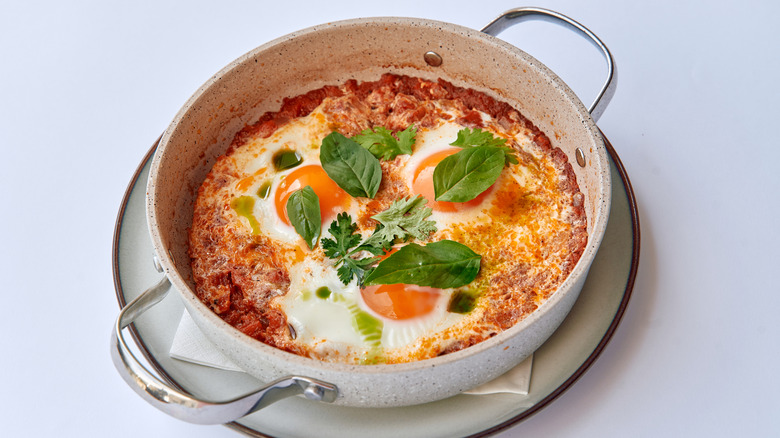 A shakshuka, made with eggs, tomatoes, and fresh herbs, is displayed fully-cooked in a Le Creuset dish
