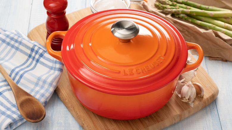 An orange Le Creuset Dutch oven on a wooden board, with seasonal items and seasonings arranged around it
