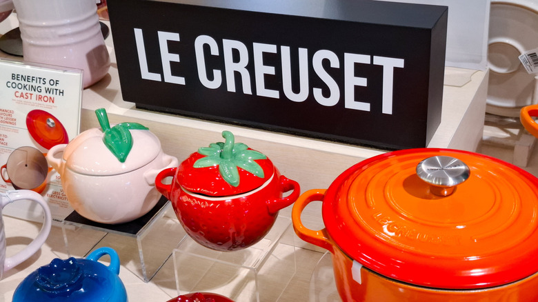 A selection of Le Creuset pots, including novelty, smaller, fruit-shaped pots, below a Le Creuset sign