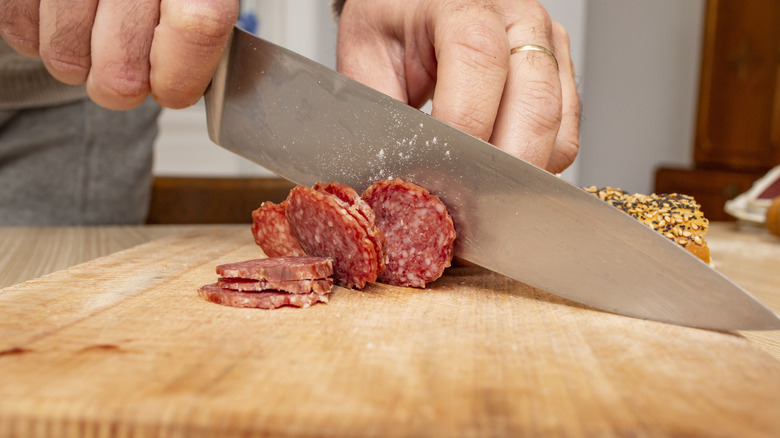 Close up of hands using large chef's knife to slice salami