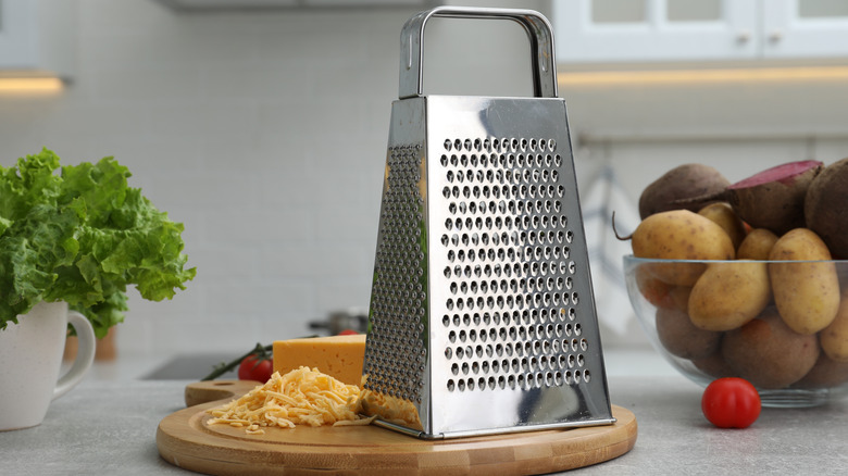 Box grater on board with shredded cheese, bowl of potatoes