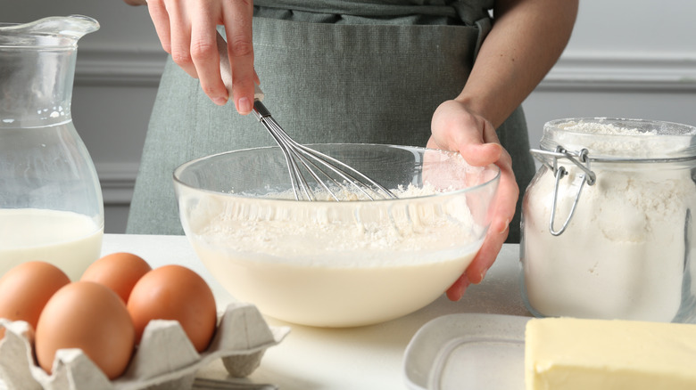 Hands whisk batter in clear mixing bowl with eggs, flour, butter nearby