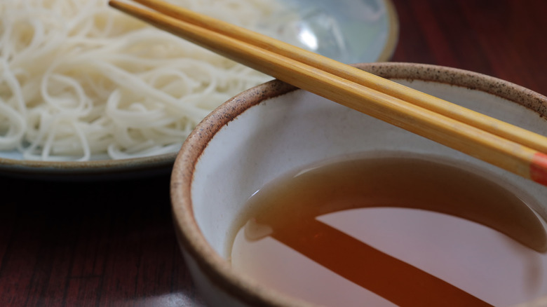 A bowl of cold noodles is seen with a bowl of mentsuyu, with a pair of chopsticks balancing on the sauce bowl
