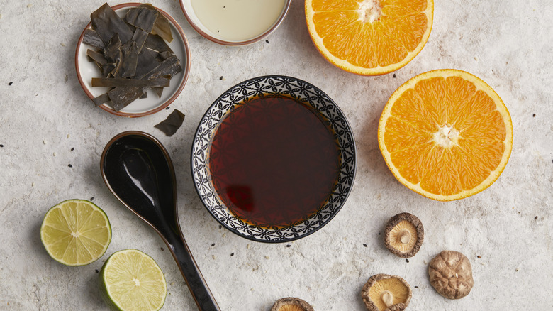 A bowl of ponzu, with various ingredients around it, like orange slices, lime halves, dried mushrooms, and kombu