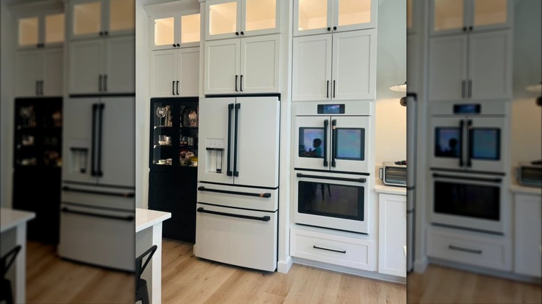 A white refrigerator and wall ovens with black handles in a white kitchen