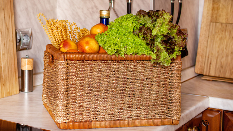 A large basket full of fruits, veggies, pasta, and a bottle on a countertop