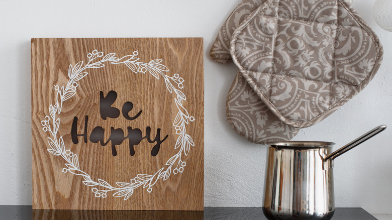 A decorative piece of wood with the words "Be Happy" leaning against the wall behind a cooktop and next to a pot and hanging potholders