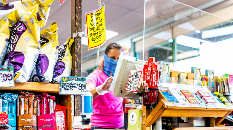 A masked a Trader Joe's cashier works behind a face shield with a sign encouraging social distancing.