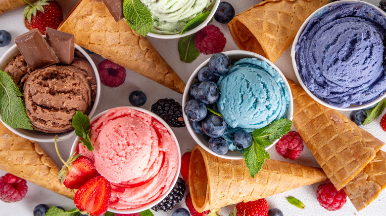 Selection of colorful scoops of ice cream in cups, surrounded by cones, berries, and other ingredients
