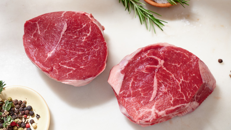 Two pieces of raw chuck tender steaks on a white surface with spices and herbs