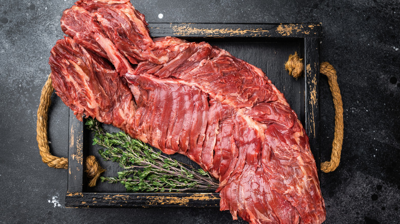 A whole hanger steak on a black surface with fresh herbs
