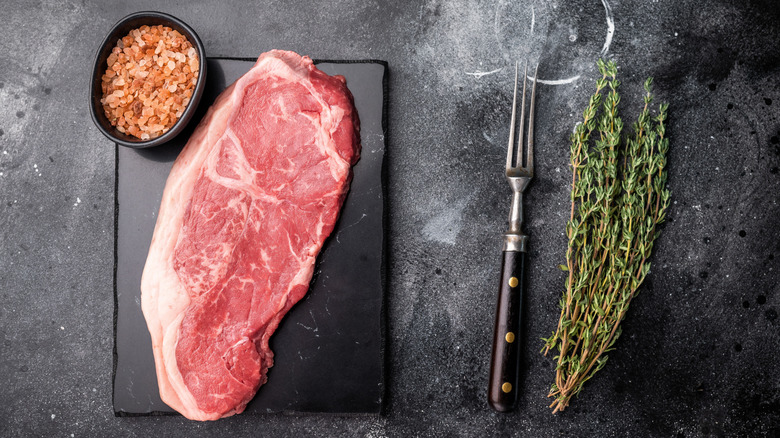 A NY strip steak on a black countertop with a fork, salt, and herbs