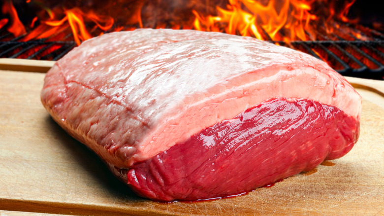 A whole picanha steak on a wooden cutting board in front of a grill